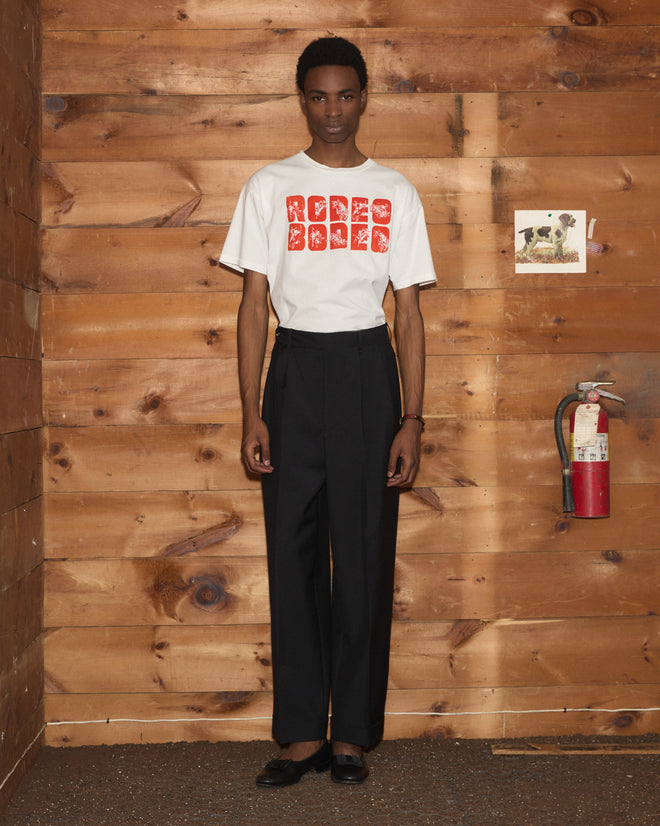 A person in a white Rodeo Bodeo Tee and black pants stands indoors by wood walls and a fire extinguisher. This vintage-inspired tee is crafted from soft 100% cotton for effortless comfort and style.