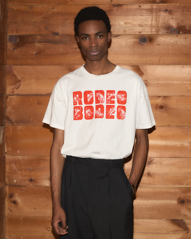 A young man wearing a white Rodeo Bodeo Tee made from 100% cotton and black pants stands by a wooden wall, looking at the camera. The Rodeo Bodeo Tee brings a vintage rodeo vibe to his laid-back style.