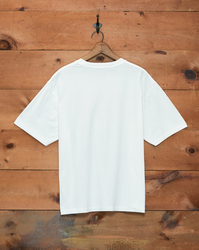 The Rodeo Bodeo Tee, crafted from 100% cotton in classic white, hangs on a wooden hanger set against a rustic wooden wall background.