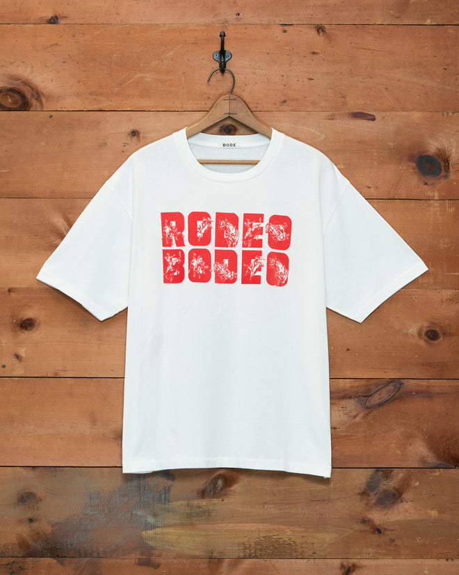 The Rodeo Bodeo Tee features bold red “RODEO” text on a white shirt, hanging on a wooden wall—a classic vintage-inspired piece crafted from soft 100% cotton. Perfect for fans of Rodeo Bodeo style.