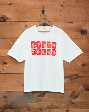 The Rodeo Bodeo Tee features bold red “RODEO” text on a white shirt, hanging on a wooden wall—a classic vintage-inspired piece crafted from soft 100% cotton. Perfect for fans of Rodeo Bodeo style.