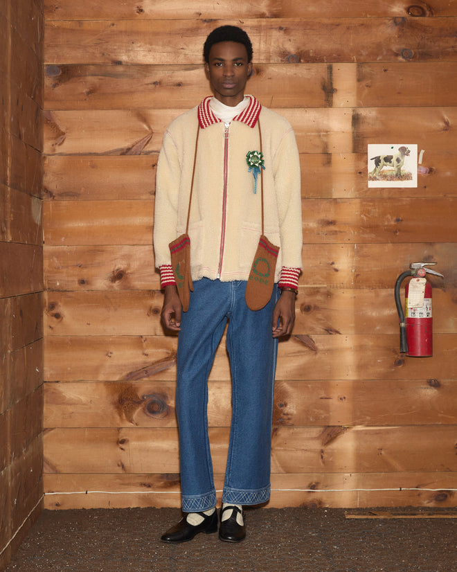 Person in a cream sherpa Woolly Warmup Jacket with a red striped collar, brown mittens, and blue jeans.