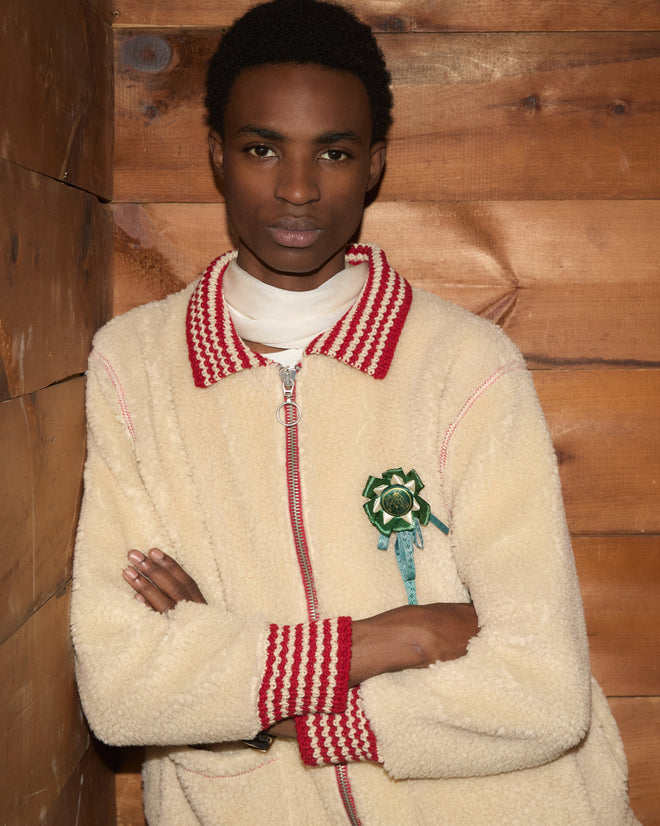 A person wears a cream-colored Woolly Warmup Jacket with red and white striped trim and a small horse ribbon pin.