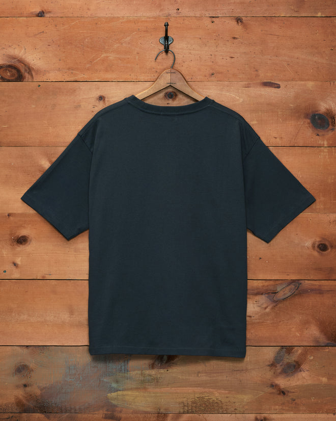A Horseshoe Stud Pocket Tee in plain black hangs on a hook against a wooden wall, shown from the back.