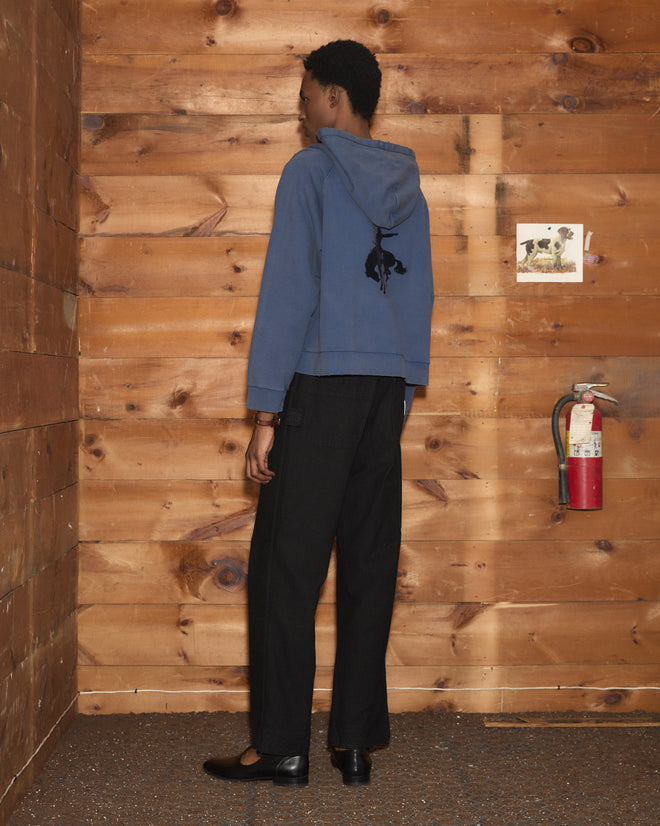 Person in a washed blue Roughrider Hoodie with a rodeo rider silhouette on the back and black trousers.