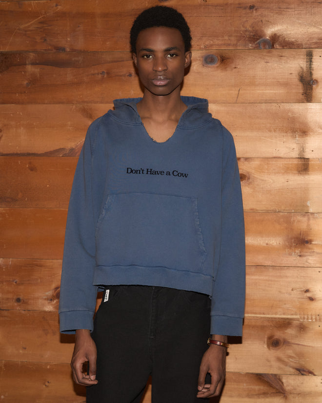 A man wears a washed blue Roughrider Hoodie with a distressed neckline and “Don’t Have a Cow” text on the chest.