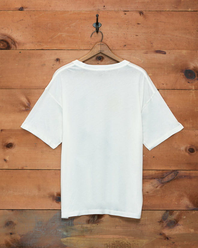The Cheval Tee, crafted from 100% cotton, hangs on a wooden hanger against a wooden wall background.