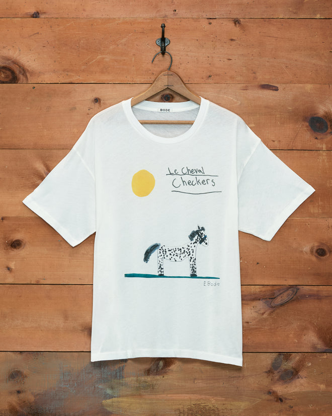 The Cheval Tee, crafted from 100% cotton for lasting comfort and charm, features a hand-drawn horse, a yellow sun, and “le cheval Checkers” text on white fabric. This childhood drawing-inspired t-shirt is pictured hanging on a wooden wall.