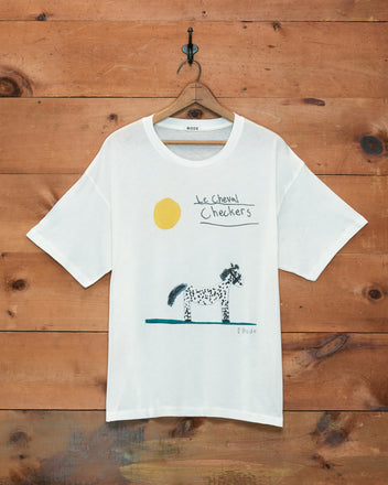 The Cheval Tee, crafted from 100% cotton for lasting comfort and charm, features a hand-drawn horse, a yellow sun, and “le cheval Checkers” text on white fabric. This childhood drawing-inspired t-shirt is pictured hanging on a wooden wall.