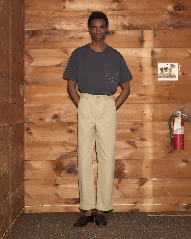 A person wearing the Horseshoe Stud Pocket Tee in gray and beige pants stands indoors by a wooden wall and a red fire extinguisher.