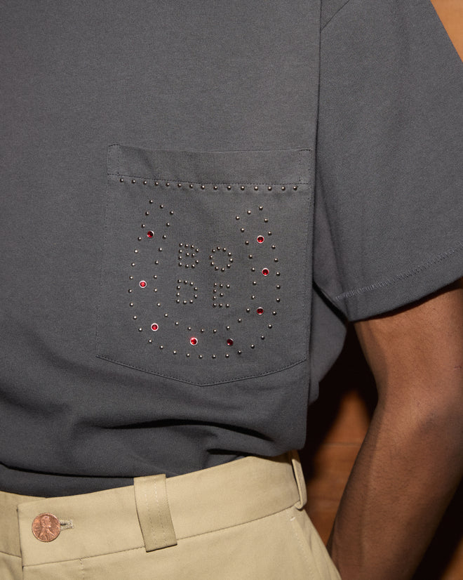 Wearing the Horseshoe Stud Pocket Tee—a gray shirt featuring “BODE” in silver studs forming a subtle horseshoe pattern on the pocket—paired with beige pants.
