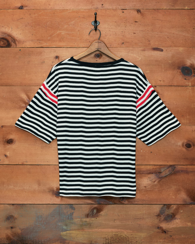 Rib Stripe Training Shirt featuring black and white stripes with bold red sleeve accents, inspired by classic 1980s jerseys, displayed on a hanger against a wooden wall.