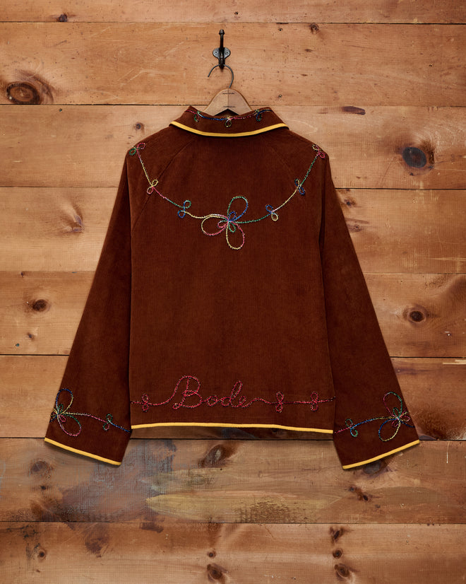 Cactus Couching Jacket—a brown corduroy piece with colorful embroidered designs and “Bode” text, featuring intricate Western chain-stitching, hanging on a wooden wall.