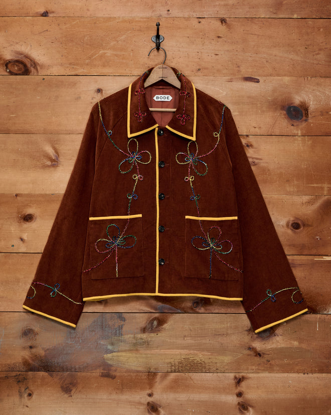 Cactus Couching Jacket—brown corduroy with yellow trim and Western chain-stitching—displayed on a hanger against a wooden wall.