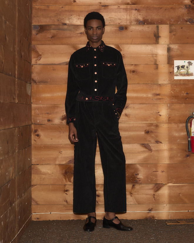 A person wearing a black Corduroy Copper Spur Jacket and matching trousers with red stitching, paired with Mary Janes.
