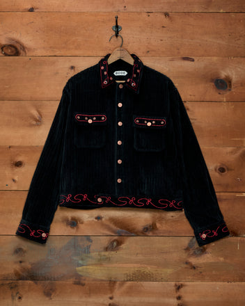 Corduroy Copper Spur Jacket: black wide-wale corduroy workwear jacket with red embroidery and copper buttons on wood.