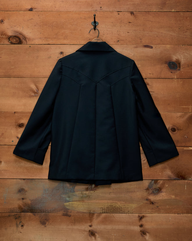 Black elongated Sonora Mohair Jacket with a pleated back and fabric-covered buttons hanging on a wooden wall.