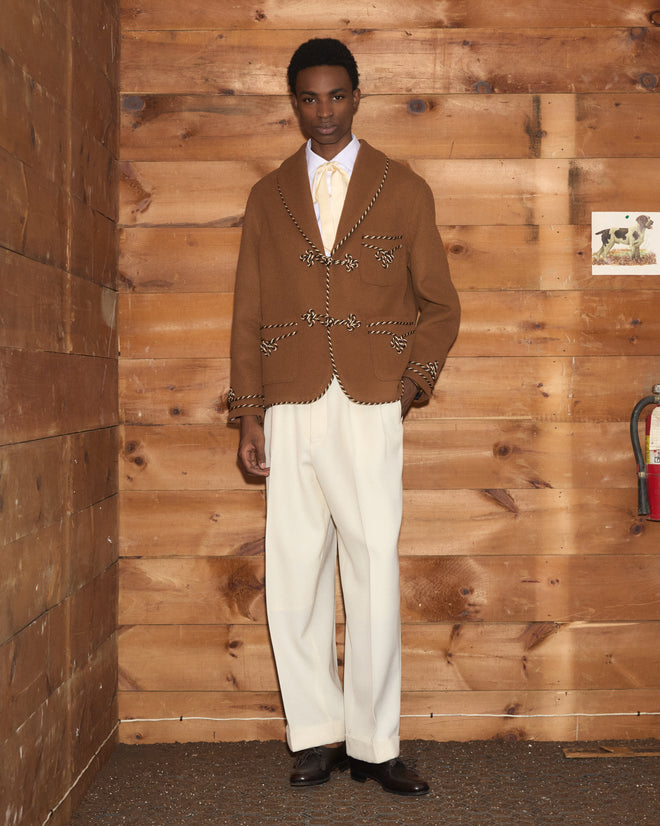 A man wears a brown velvet Bradford Smoking Jacket with rope toggles and white trousers.