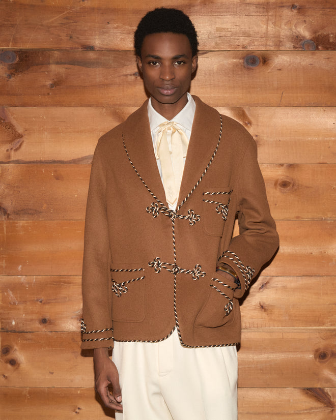 A person wears a brown velvet Bradford Smoking Jacket with braided rope toggles over a white shirt and cream trousers.