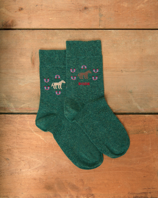 Horseshoe Socks in green, featuring a white dinosaur with purple spots and the word “Roar” embroidered, displayed on a wooden surface.
