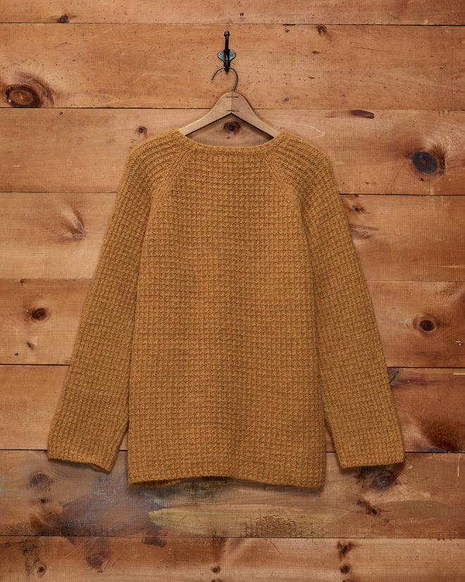 An orange Show Sweater with a textured knit hangs on a wooden hanger against a rustic wooden wall background, evoking the classic style often seen at an equestrian show.