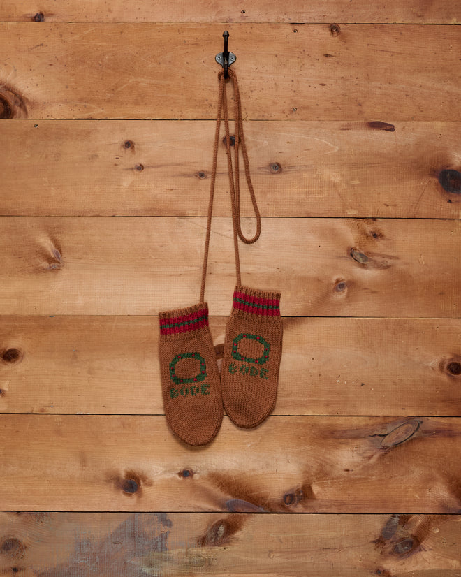 A pair of Holly Stripe Mittens, children’s brown mittens with green designs and a subtle wreath pattern, hangs on a hook against a wooden wall.