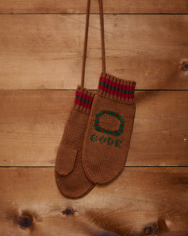 The Holly Stripe Mittens, featuring a brown knit design with red and green wreath pattern details, hang on a wooden wall.