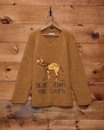 Show Sweater - Orange, a cozy merino knit featuring a charming camel graphic and the phrase “BLUE JEANS AND CHIPS,” displayed against a wooden wall—an ideal pick for subtle equestrian-inspired style.