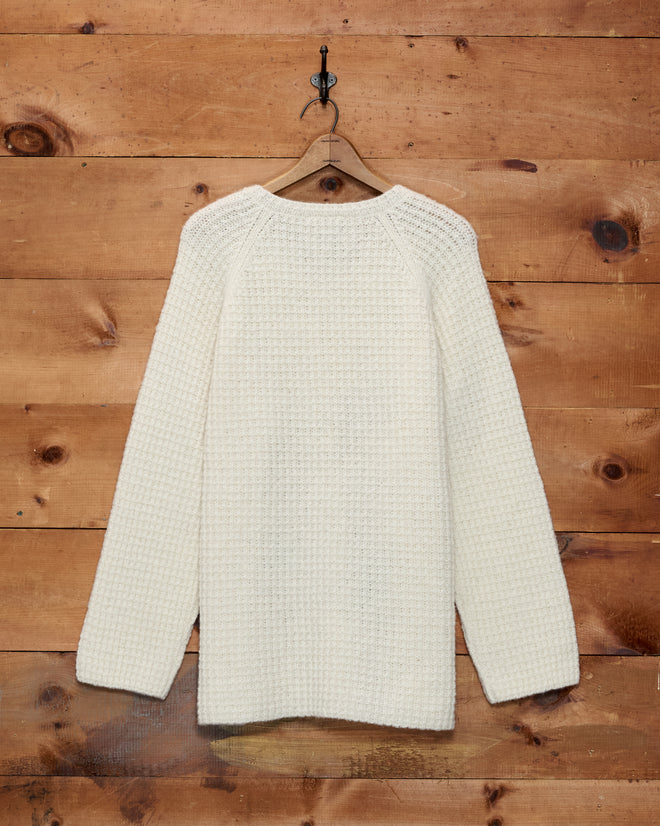 The Show Sweater - Ivory hangs on a wooden hanger against a wooden wall background, evoking the refined style of an equestrian show.