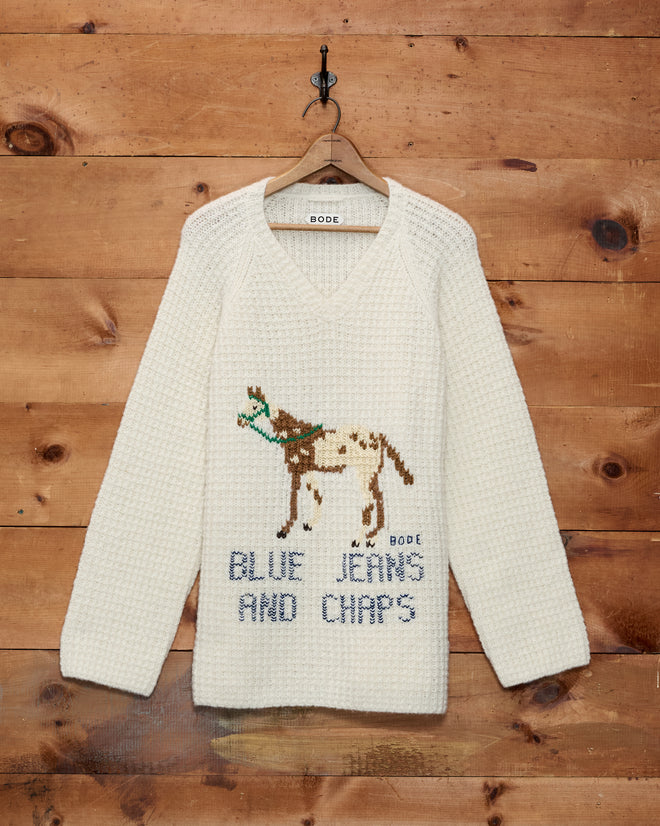 Show Sweater - Ivory with a knit horse graphic inspired by the Pony of the Americas and the words “BLUE JEANS AND CHAPS,” hanging on a wooden wall.