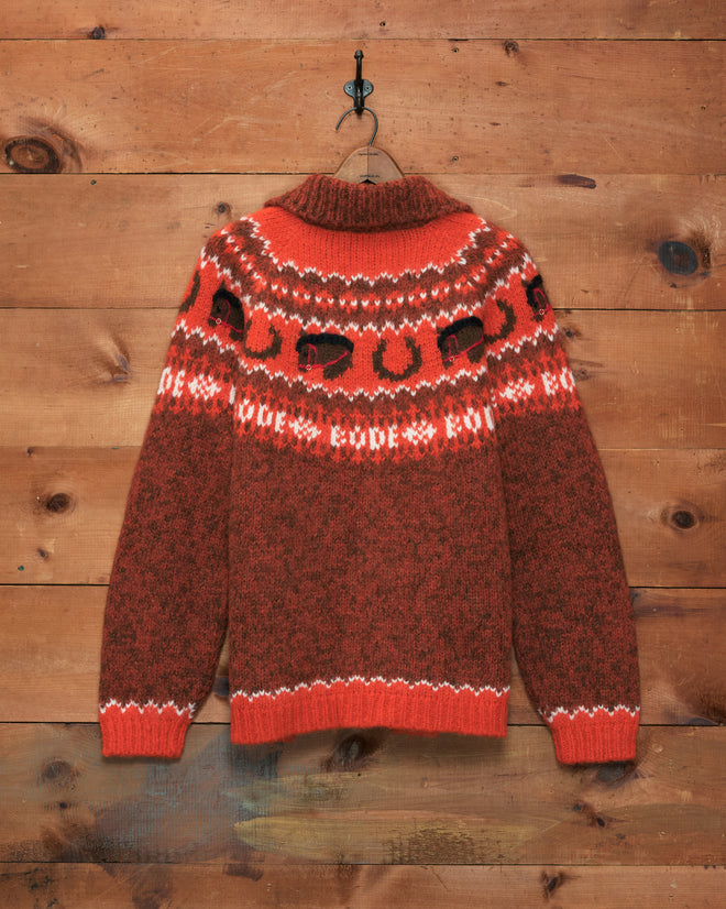 The Corral Cardigan, a red and brown knit sweater featuring white and black patterns and crafted in Peru, hangs on a wooden wall.