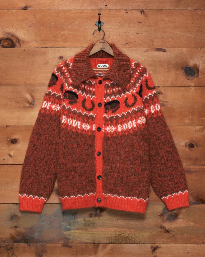 The Corral Cardigan, featuring a red and brown pattern and crafted from alpaca in Peru, hangs on a wooden wall hook.