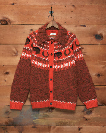 The Corral Cardigan, featuring a red and brown pattern and crafted from alpaca in Peru, hangs on a wooden wall hook.