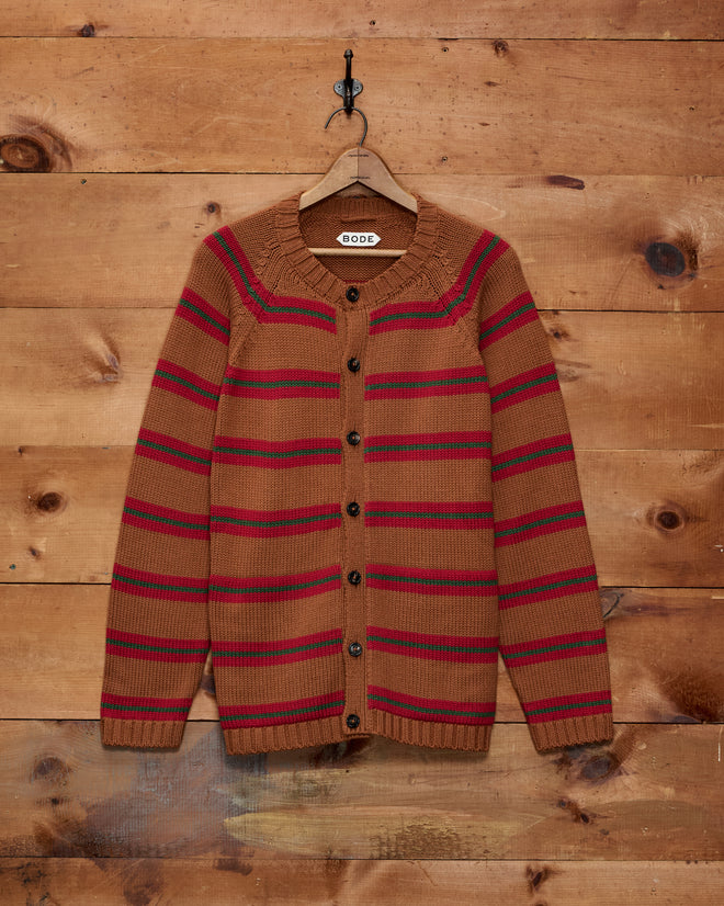 The Holly Stripe Cardigan, crafted from brown cotton wool with red and green stripes, hangs on a wooden wall.
