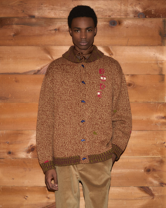 A person wearing the Farrier Cardigan paired with tan pants stands in front of a wooden wall, looking at the camera.