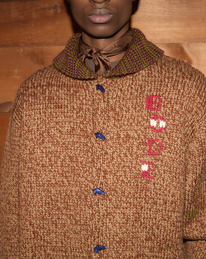 A person wearing the Farrier Cardigan—a chunky brown wool knitwear sweater featuring horsehead buttons and a patterned collar—paired with a brown scarf tied at the neck.
