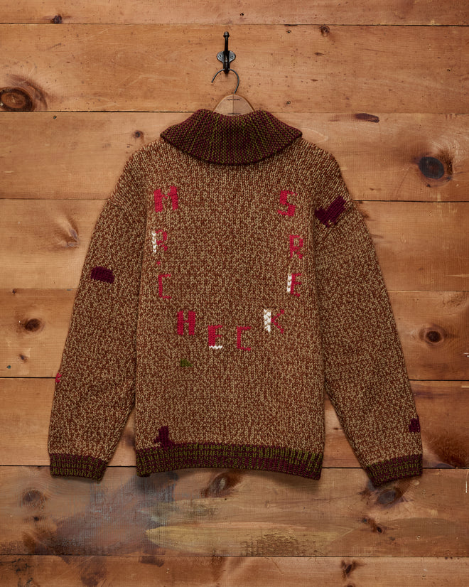 The Farrier Cardigan—a brown wool knitwear sweater adorned with scattered red and white letters—hangs on a wooden wall by a metal hook, showcasing its heavyweight construction and distinctive horsehead buttons.