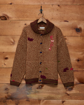 The Farrier Cardigan, a brown wool knitwear sweater featuring animal patterns, hangs on a wooden wall and is displayed on a hanger with a label.