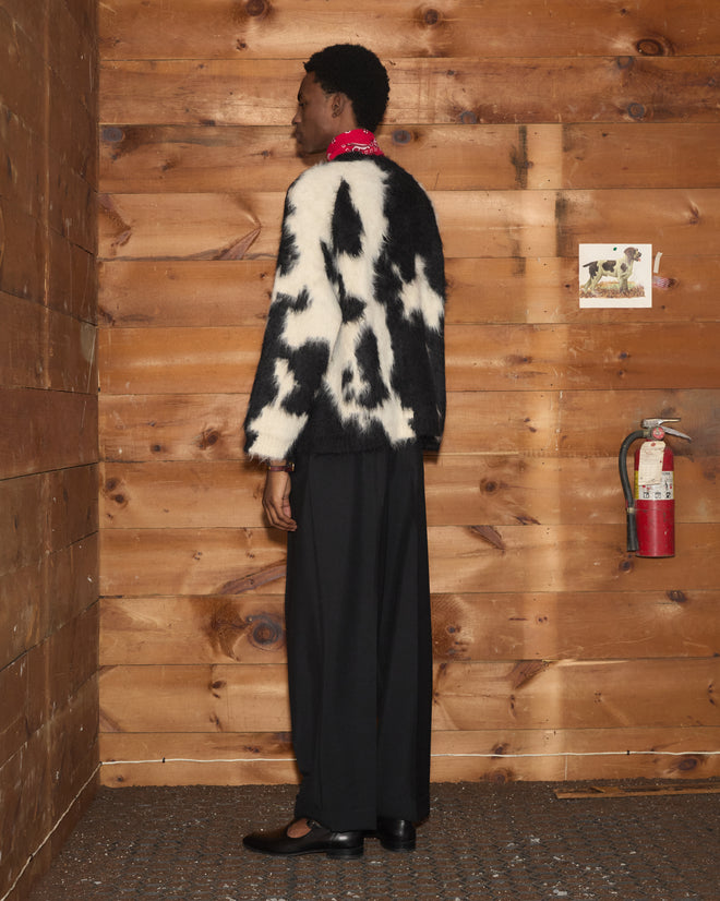 A person in the Holstein Sweater - Black/White, black pants, and a red scarf stands by a fire extinguisher against a wood wall.