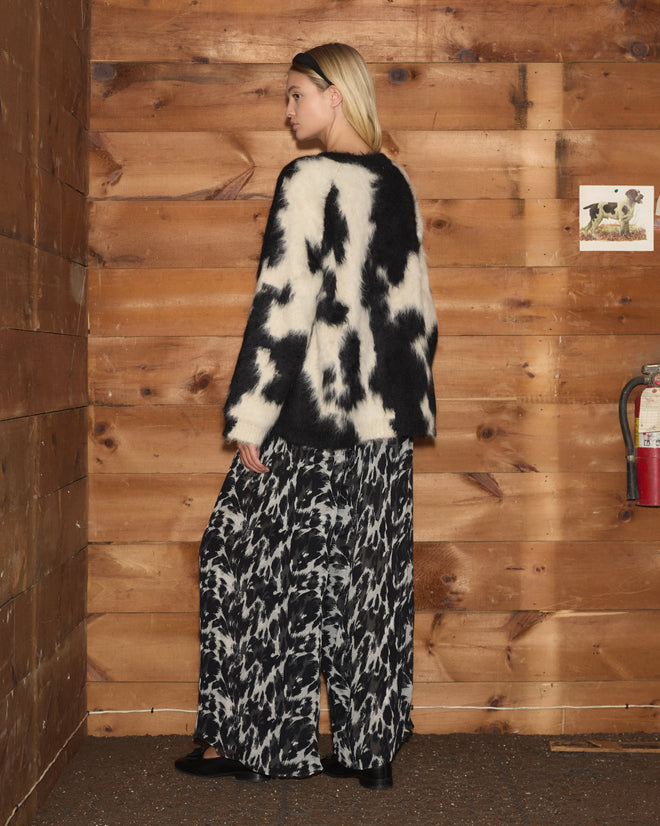 A woman wearing the Holstein Sweater in Black/White and patterned pants stands in a wooden room, facing away from the camera.