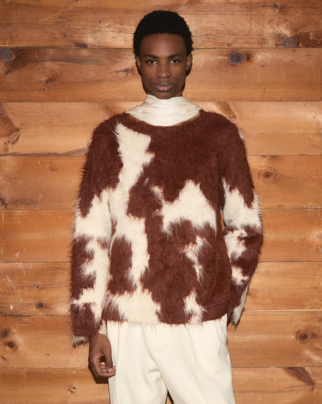 A person wearing the Holstein Sweater in Brown/White, paired with cream pants, stands against a wooden wall.