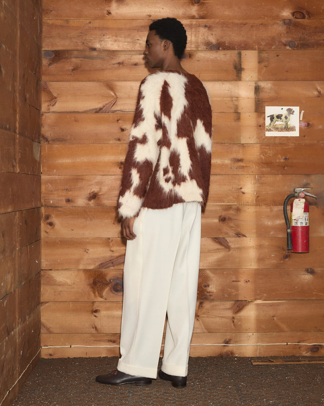 A person wearing the Holstein Sweater - Brown/White and white pants stands sideways against a wooden wall with a fire extinguisher.