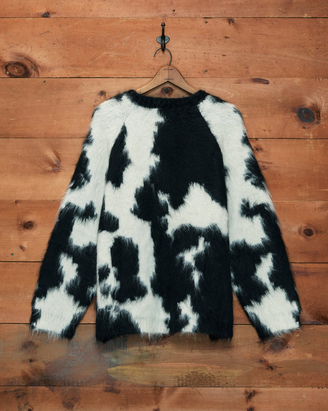 The Holstein Sweater in Black/White, crafted from a soft brushed alpaca knitwear blend, hangs on a wooden wall, showcasing its fuzzy black and white cow print pattern.