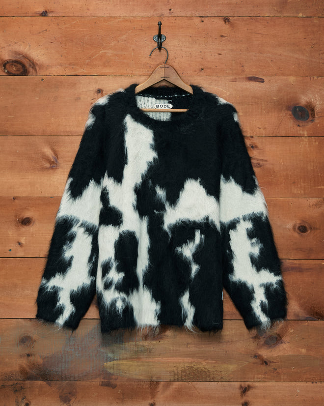 The Holstein Sweater in black and white, featuring a fuzzy brushed alpaca knit with a cow print pattern, is displayed hanging on a wooden wall.