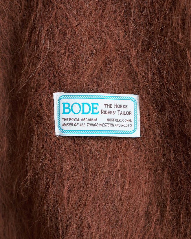 A white BODE label sewn onto soft, fuzzy brown-and-white knit fabric, reading “THE HORSE RIDERS’ TAILOR” and other text, is attached to the Holstein Sweater – Brown/White.