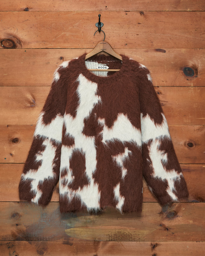 The Holstein Sweater in Brown/White hangs on a wooden wall, its soft knitwear texture adding a cozy charm to the scene.