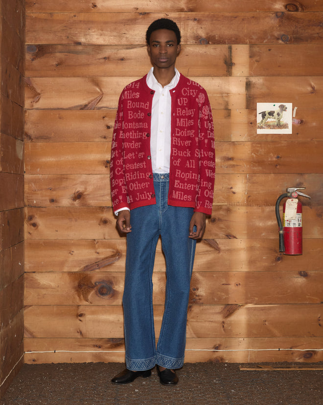 A person stands indoors against a wood wall, wearing the Beaded Round-Up Cardigan in red over a white shirt and blue jeans.