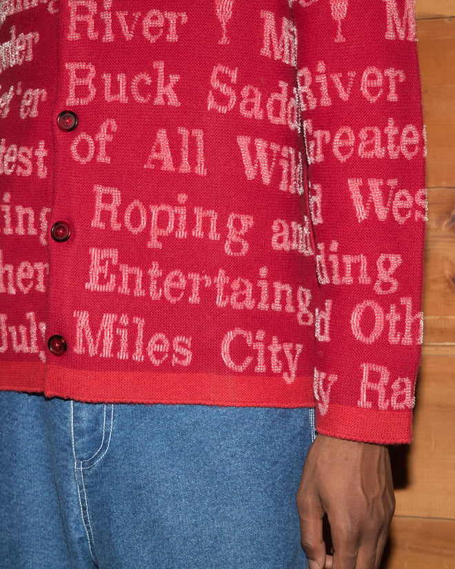 A person wearing the Beaded Round-Up Cardigan—a red wool-cashmere blend sweater with white text—paired with blue jeans stands near a wooden wall, capturing the classic rodeo style inspired by the Miles City Round-Up.