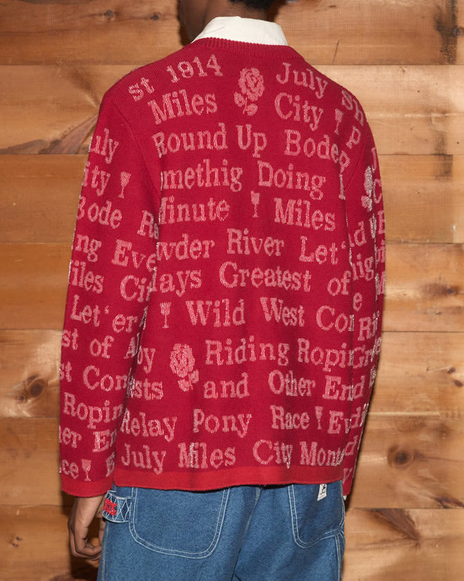 A person wearing the Beaded Round-Up Cardigan—a red wool cashmere sweater with white text—paired with blue jeans, stands in front of a wooden wall, evoking the classic rodeo style associated with the Miles City Round-Up.