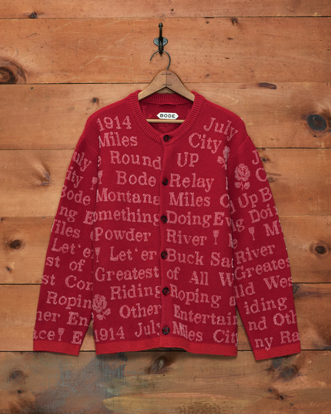Beaded Round-Up Cardigan in red with scattered light pink text, hanging on a wooden wall on a hanger with a hook.